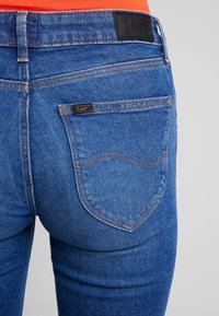 Modré džíny z denimu s detailním záběrem na zadní kapsu, která obsahuje koženou nášivku s logem a kontrastní prošívání.