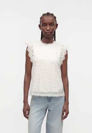 Γυναίκα με πλεξούδες φοράει λευκό δαντελωτό τοπ χωρίς μανίκια με κυματιστές άκρες και ανοιχτό μπλε τζιν, στέκεται μπροστά σε ένα απλό φόντο.