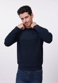 Sweat-shirt en maille bleu marine avec texture côtelée, col rond et manches longues, arborant un motif horizontal sur la poitrine.