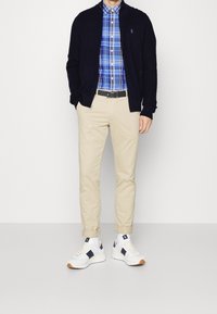 Laivastonsininen neuletakki, sininen ruudullinen paita, beiget chinot ja valkoiset tennarit laivastonsinisin yksityiskohdin. Laadukkaat materiaalit ja räätälöity istuvuus.