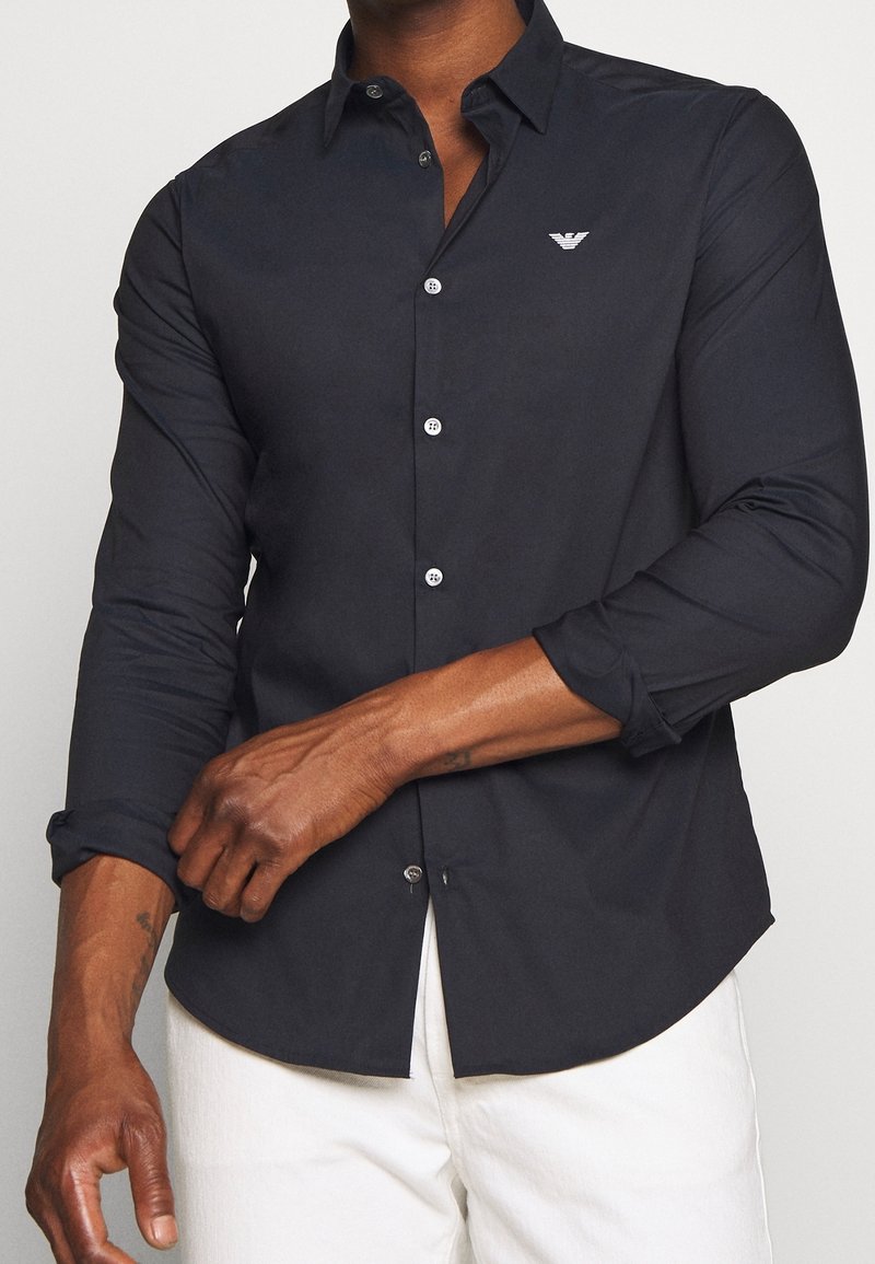 Muž si vyhrnuje rukávy černé přiléhavé košile na knoflíky s bílým znakem, v kombinaci s bílými kalhotami na jednobarevném pozadí.