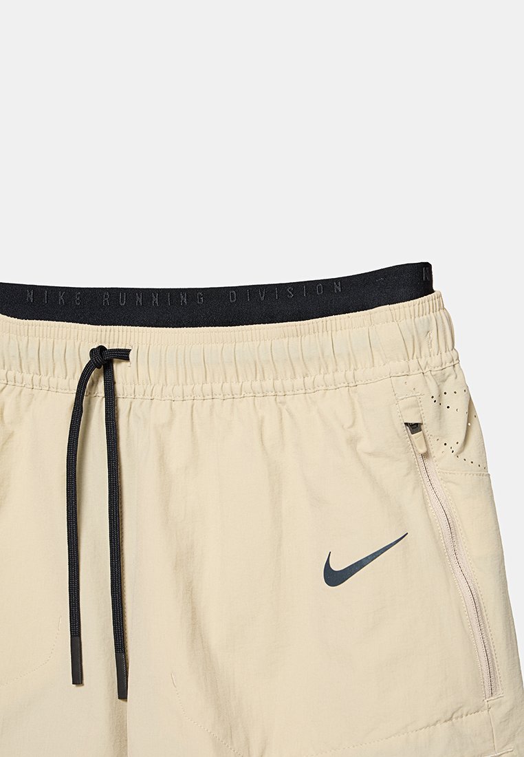 Shorts de correr beige claro con cinturilla elástica, cordón negro, bolsillo con cremallera lateral y logo de Nike en gris oscuro. Presenta orificios de ventilación.