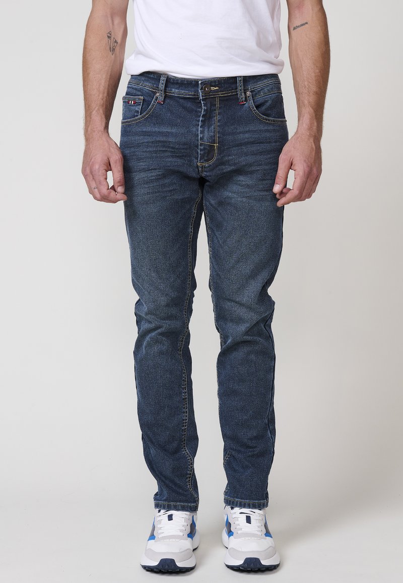 Jeans de mezclilla azul oscuro con corte slim. Presentan costuras amarillas, diseño de cinco bolsillos y cierre de botón. Combinados con zapatillas blancas y azules.