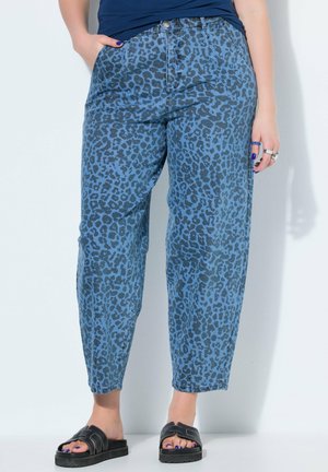 Frau trägt blaue Jeans mit Leopardenmuster, schwarze Slide-Sandalen und dunkelblaues Oberteil, steht mit den Händen nahe den Taschen vor weißem Hintergrund.