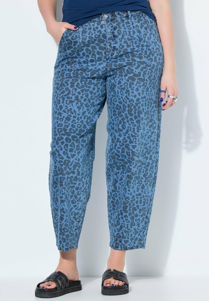 Mujer con vaqueros azules con estampado de leopardo, sandalias negras tipo slide y top azul oscuro, de pie con las manos cerca de los bolsillos sobre fondo blanco.