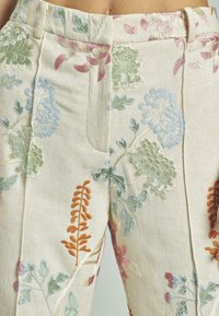 Pantaloni in lino ricamati con uno sfondo crema, caratterizzati da intricati disegni floreali in fili blu, verdi, arancioni e rosa.