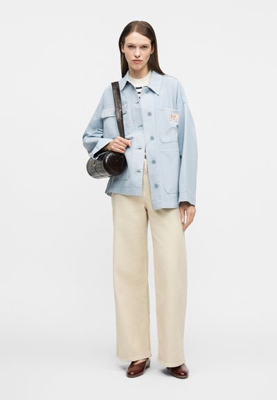 Γυναίκα που φοράει ανοιχτόχρωμο μπλε φαρδύ μπουφάν, ριγέ τοπ, μπεζ παντελόνι με φαρδιά πόδια, καφέ παπούτσια και κρατά μια μαύρη στρογγυλή τσάντα χιαστί.