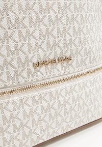 Gros plan d'un sac beige Michael Kors avec un motif répétitif de logo MK, une fermeture éclair dorée et une plaque avec le nom de la marque.