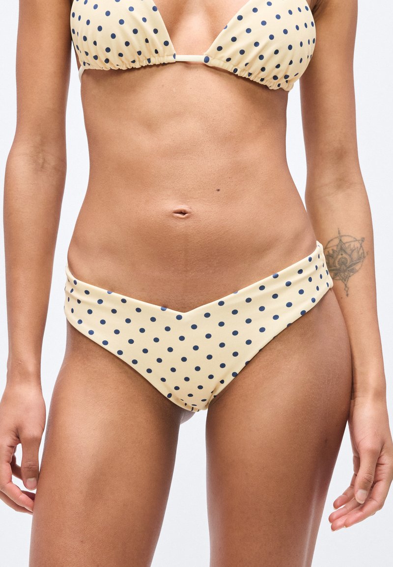 Γυναίκα που φοράει μπεζ μπικίνι με μπλε πουά σημεία· τατουάζ από ρόδα του αέρα στο αριστερό πήχη ορατό.