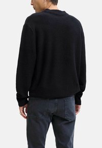 Maglione nero in pile con superficie strutturata, spalle scese e polsini a coste. Abbinato a jeans grigio scuro, presenta un dettaglio posteriore visibile.