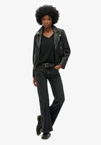 Zwarte leren motorjack, kortbermde zwarte shirt, donkere flared jeans en zwarte laarzen met gestructureerde zolen. Model staat met de handen op de heupen.