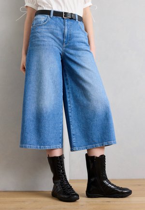 Person som har på seg vide, croppede blå jeans med svart belte og svarte snørestøvletter, står på tregulv.