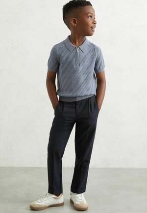 Polo-shirt en tricot bleu clair avec un col zippé, associé à un pantalon noir marine et des baskets blanches avec des accents beige.