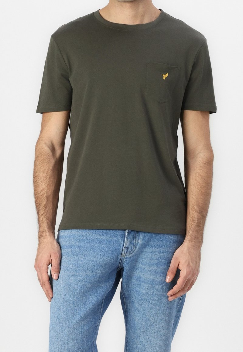 Homme portant un t-shirt à manches courtes vert olive avec une poche poitrine et un petit logo d'oiseau jaune, associé à un jean bleu clair.