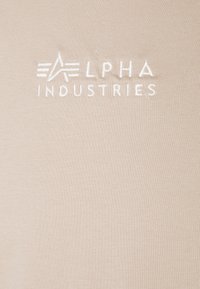 Camiseta beige claro hecha de tela suave, con el logotipo bordado en blanco "ALPHA INDUSTRIES" y una textura elegante y lisa.