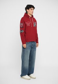 Rød hættebluse med broderede detaljer, der inkluderer logoer og mærker. Sammen med løse blå denimjeans og hvide sneakers.