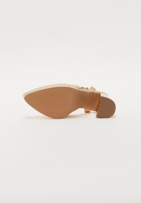 Scarpa con tacco alto color beige, con finitura liscia, punta affusolata e cinturino alla caviglia. Suola in gomma testurizzata e tacco quadrato piccolo per la stabilità.