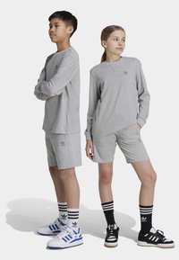 Graue Sportbekleidung besteht aus einem Langarmoberteil und Shorts, die mit dem Adidas-Logo versehen sind. Die Modelle tragen gestreifte Socken und kontrastierende Sneaker.