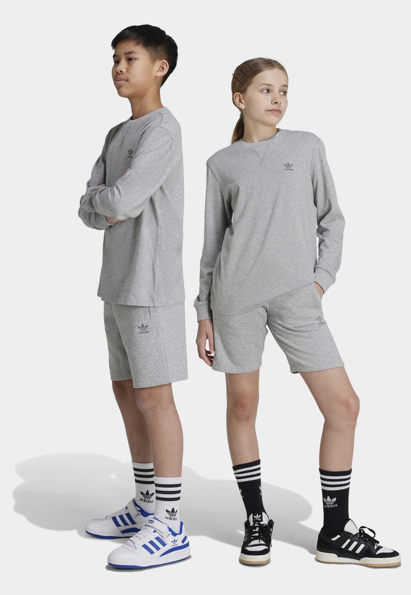 Graue Sportbekleidung besteht aus einem Langarmoberteil und Shorts, die mit dem Adidas-Logo versehen sind. Die Modelle tragen gestreifte Socken und kontrastierende Sneaker.