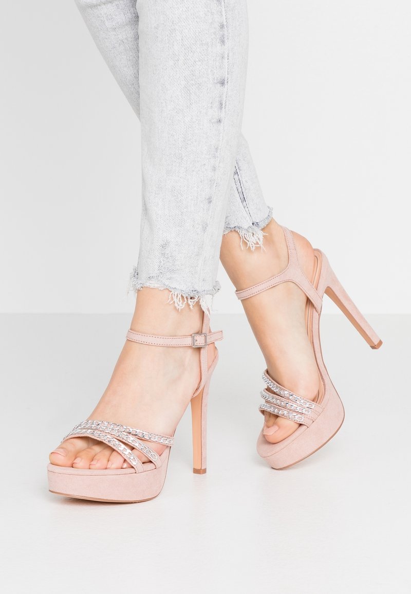 Personne portant des sandales à plateforme à talons hauts roses clairs avec des brides en strass et un jean bleu clair à ourlet effiloché sur un sol blanc.