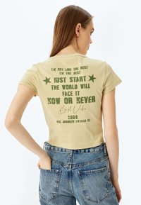 Lysegul kortærmet t-shirt med grøn trykt tekst og stjerner. Teksten indeholder motiverende sætninger, og designet inkluderer en grafik fra 2008.