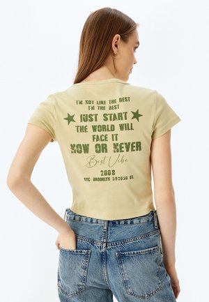 Lichtgele cropped t-shirt met groene bedrukte tekst en sterren. De tekst bevat motiverende zinnen en het ontwerp heeft een grafische afbeelding uit 2008.