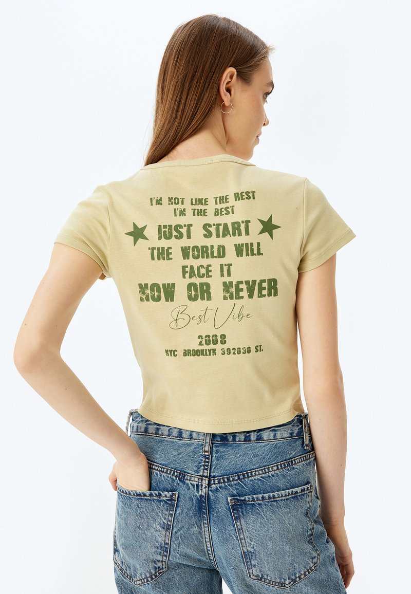 Lysegul kortærmet t-shirt med grøn trykt tekst og stjerner. Teksten indeholder motiverende sætninger, og designet inkluderer en grafik fra 2008.