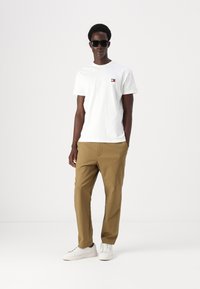 Tricou alb din bumbac cu un mic logo roșu și albastru pe piept, asociat cu pantaloni khaki tapered și adidași albi. Trăsături notabile includ ochelari de soare.