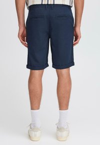 Marineblå shorts med oppbrettede ermer, to baklommer, glatt tekstur og et rent design, kombinert med lyse sko og hvite sokker.
