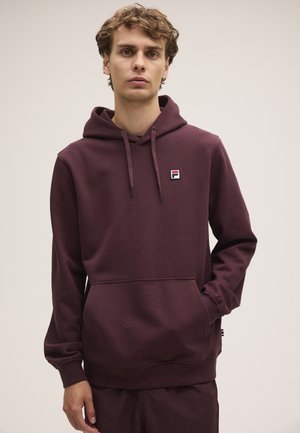 Giovane uomo che indossa una felpa con cappuccio bordeaux scuro con un piccolo logo sul petto e la mano sinistra nella tasca anteriore.