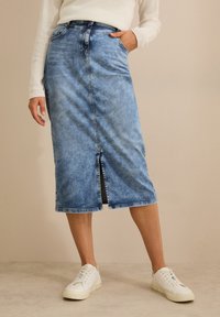 Midi-Jeansrock in Hellblau, mit einem Schlitz vorne, Fünf-Taschen-Design und subtilen Ausbleichungen. Kombiniert mit weißen Sneakern.