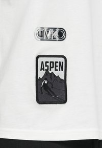 Balta T-krekls ar sudrabotu logo uzlīmi un melnu izšūtu uzlīmi ar uzrakstu "ASPEN", dizainā attēlots slēpotājs un kalni.