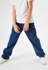 Vaqueros de mezclilla azul con un corte holgado, piernas rectas y detalles de costura visibles. Combinados con zapatillas blancas sobre un fondo neutro.