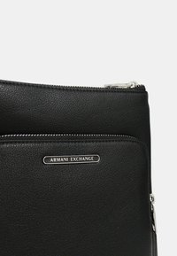 Borsa in pelle nera con texture e hardware argentato. Presenta una tasca con zip e un logo Armani Exchange in rilievo sul davanti.