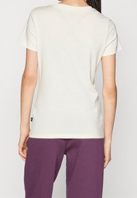 Person som bär en kortärmad krämfärgad t-shirt och lila byxor, sedd bakifrån, med flätat hår.