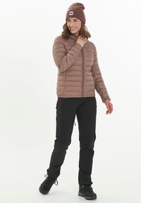 Lichtbruin pufferjack met horizontale stiksel, zwarte broek met ritsen en donkere laarzen, aangevuld met een bruine beanie met een logo.