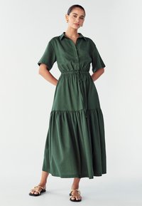 Abito verde con bottoni, maniche corte, vita elastica e gonna a strati. Realizzato in tessuto texturizzato con una vestibilità comoda. Sandali neri con dettagli dorati.