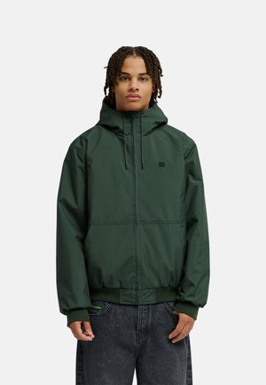 Junge Person mit geflochtenem Haar, die eine dunkelgrüne Kapuzenjacke und dunkle Jeans trägt, steht vor einem schlichten hellen Hintergrund.
