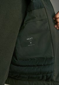 Oliven grøn jakke med en tekstureret strikoverflade, glat inderside og en sidelomme. Etiket læser "NEXT," størrelse "M," fremstillet i Kina.