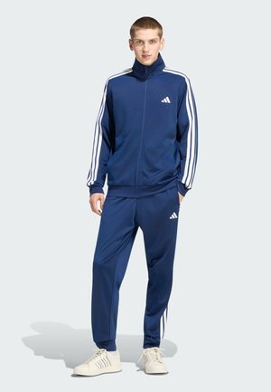 Junger Mann steht in einem marineblauen Trainingsanzug mit weißen Streifen und Logo, kombiniert mit weißen Sneakers, vor einem einfarbigen hellen Hintergrund.
