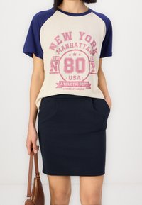 Une personne portant un T-shirt beige et marine avec l'impression "New York Manhattan" et une jupe marine, tenant un sac à main marron à ses côtés.