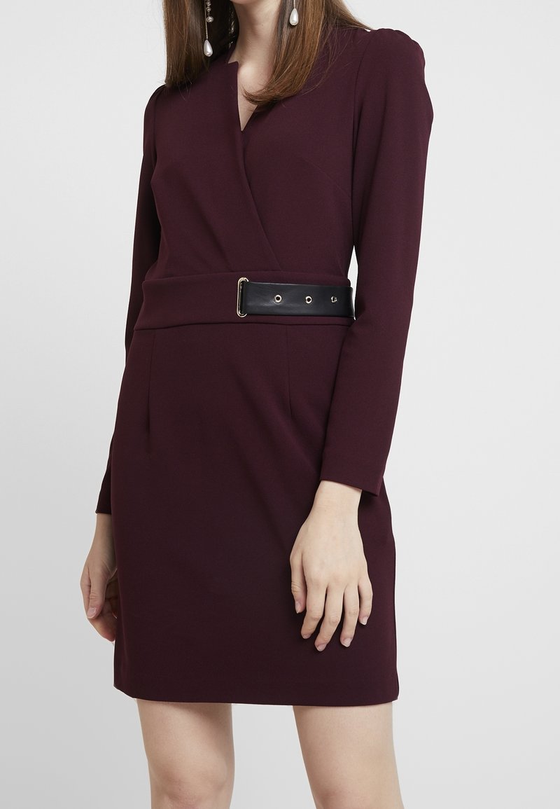 Femme portant une robe portefeuille à manches longues de couleur violet foncé avec une ceinture noire et des boucles d'oreilles en perles pendantes, posant devant un fond gris uni.