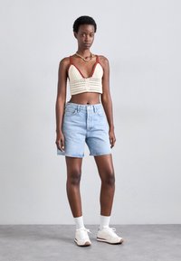Bralette all'uncinetto in crema con dettagli rossi, abbinato a pantaloni corti in denim azzurri. Il modello indossa sneakers bianche e calzini di lunghezza media.