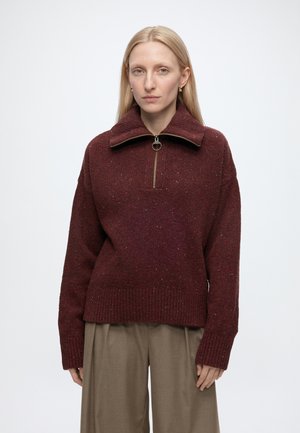 Frau mit langen blonden Haaren, die einen bordeauxroten Zip-Up-Pullover und beige weite Hosen vor einem schlichten weißen Hintergrund trägt.