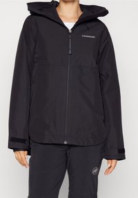 Veste de pluie noire zippée avec capuche et poignets ajustables, portée avec un pantalon noir arborant un logo mammouth sur la cuisse.