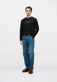 Black long-sleeve shirt with white logo on chest, paired with blue straight-leg jeans and brown leather boots, standing on a neutral background.