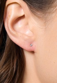 Srebrna kolczyk w kształcie serca z przezroczystym kamieniem, prezentowany na uchu modelki, z średnio brązowymi włosami i odcieniem skóry.