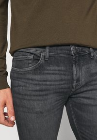 Hombre vistiendo jeans ajustados gris oscuro y un suéter de manga larga verde oliva, con la mano izquierda relajada a un lado.