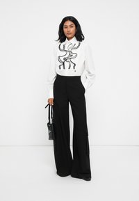 Camicia bianca con dettagli in raso neri, abbinata a pantaloni neri a vita alta e gamba larga. Il modello tiene una borsa nera. Sfondo semplice.