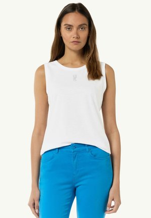 Biała bezrękawnikowa bluzka z okrągłym dekoltem i małym graficznym detalem z przodu, w połączeniu z jasnoniebieskimi spodniami. Gładka tekstura tkaniny.